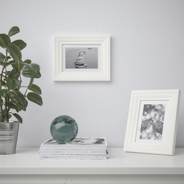 A shelf displays a plant, stack of books, and two white picture frames. One frame holds a black-and-white boat image, the other a butterfly.