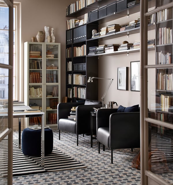 A stylish lounge with black EKERÖ armchairs, bookshelves, and a geometric rug. Large windows let in natural light.