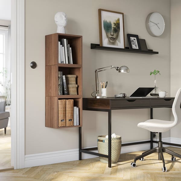Home office with white chair, black desk, wooden shelves, lamp, laptop, plants. EKET shelves hold books, baskets. Clock and art decorate wall.