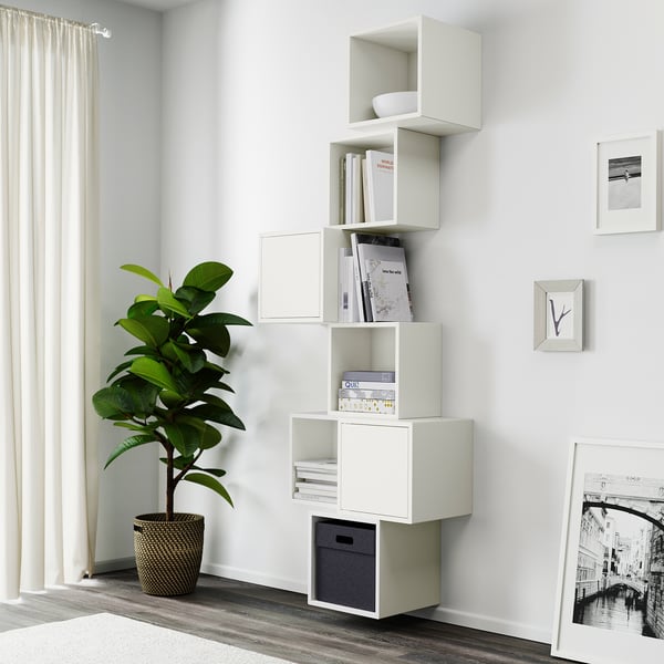 White modular wall-mounted shelves organise books and decor near a window, with a tall green plant beside them.