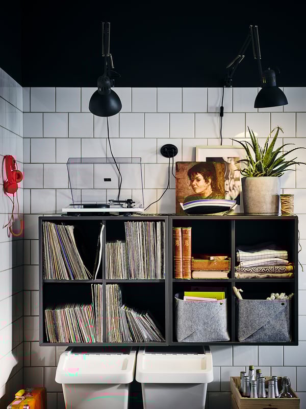 Black EKET wall-mounted storage unit with shelves and bins, holding records, books, and decor, featuring industrial lights above.
