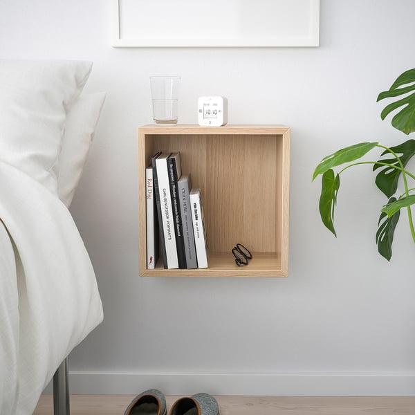 Small wooden wall shelf, light wood colour, square shape. Contains books, alarm clock, glass, and scissors. Located near bed, smaller than plant.