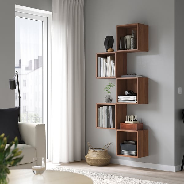 EKET wall-mounted shelves in walnut colour, holding books and decor, placed next to a window and a sofa. Its modular design maximises wall space.