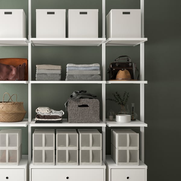 White ELVARLI wardrobe system, adjustable shelves and compartments storing towels, bags, and decor against a dark green wall.