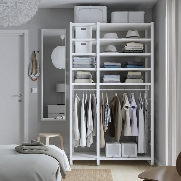 Modern bedroom with ELVARLI white storage. Corner shelving holds clothes and hats. Hangs garments below. Surrounded by bed and mirror.