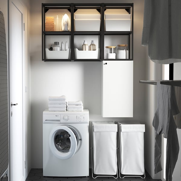 Modern laundry setup: white ENHET wall cabinet & open frame, holding towels & bottles. White washing machine fits underneath. Four laundry baskets.