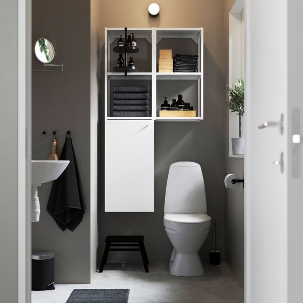 Modern bathroom with ENHET storage, sink, and toilet. Open cabinets show towels and toiletries. Round mirror and stool for functionality.