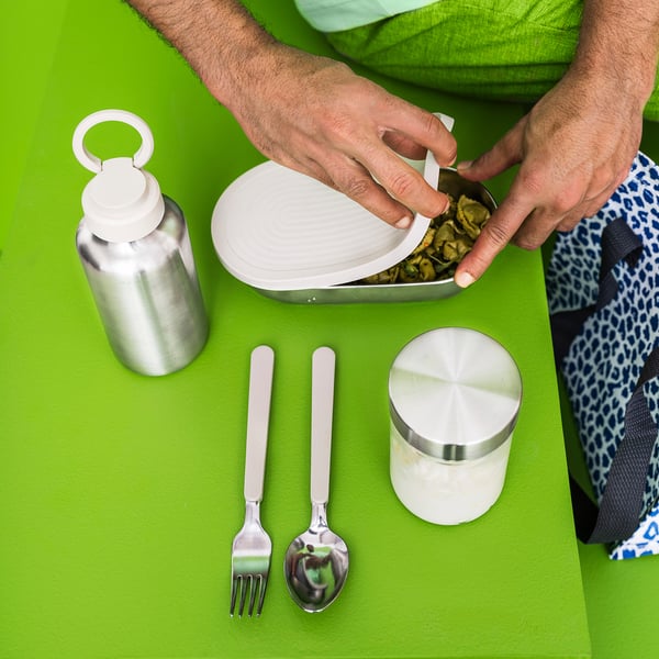 Person holding lunch box with utensils and container on bright green surface, featuring silver water bottle and stainless items.