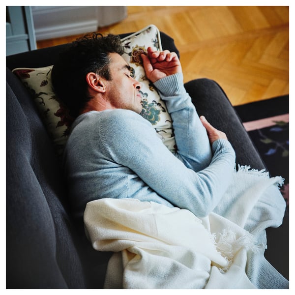 Person reclining on soft, black ESSEBODA sofa, draped in white blanket, wearing light blue sweater.