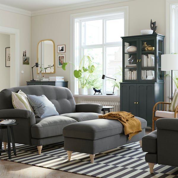 Modern living room: grey ESSEBODA sofa with tufted back, striped rug, black cabinet, plants, and gold mirror.