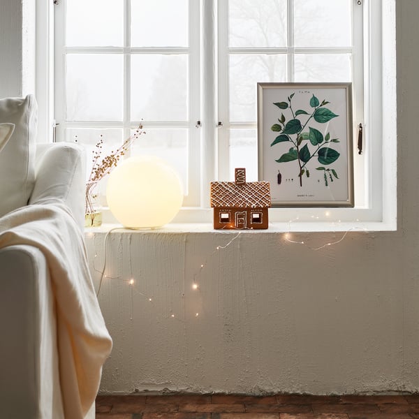Cosy living area with large white globe lamp on window sill, decorated with fairy lights and small gingerbread house.