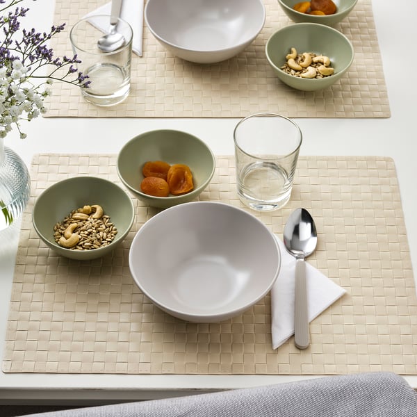Table setting with beige woven placemats, white bowls, green bowls filled with dried apricots and mixed nuts, clear glasses, silver spoon, and napkin.