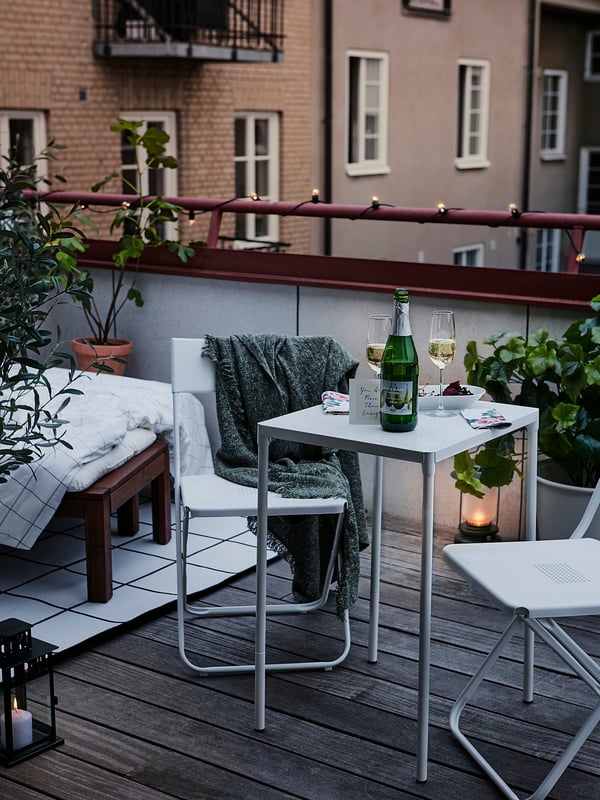 Modern balcony setting with white foldable tables and chairs, green blanket, wine glasses, candle, and plants.