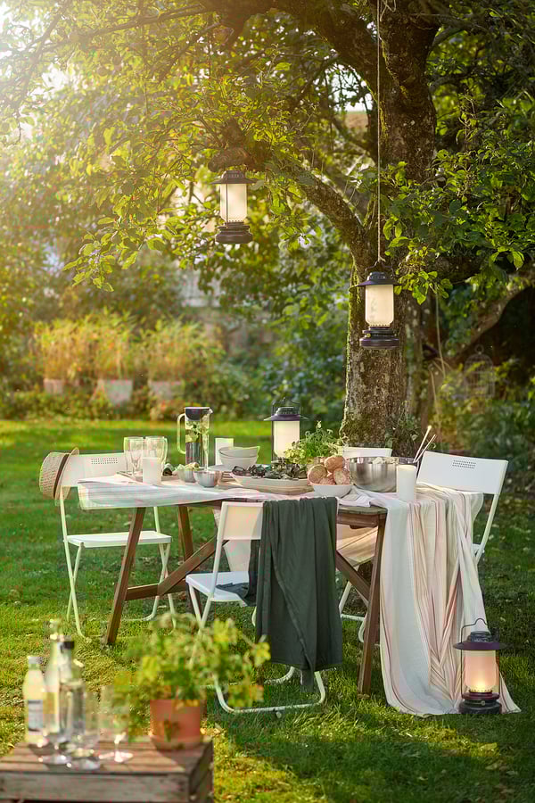 Outdoor garden party setup under tree with table, chairs, lanterns, cutlery, plates, towels, and blankets.