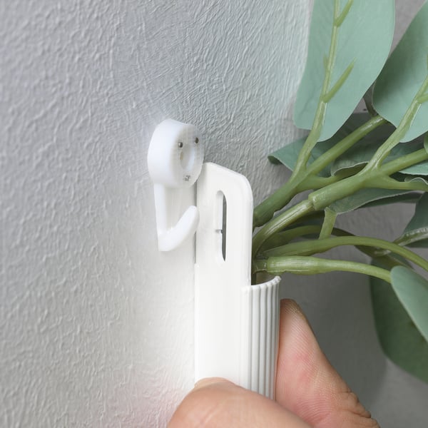 A hand instals a white wall-mounted planter for FEJKA artificial plant. The planter uses an adhesive hook, suitable for indoor/outdoor use.