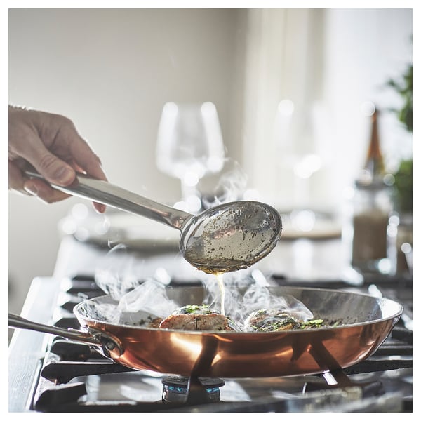Hand drizzles liquid from spoon into sizzling pan on gas stove.