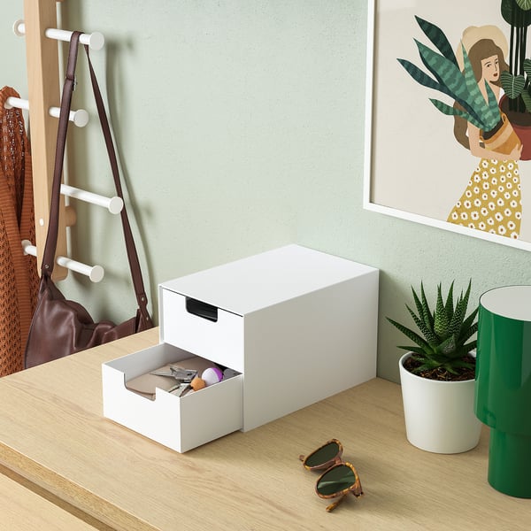 White mini storage chest with one drawer open, holding small items. Placed on a wooden surface near a coat rack and a framed poster.