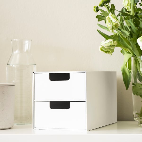 White mini storage chest with two drawers on white table, next to vases.