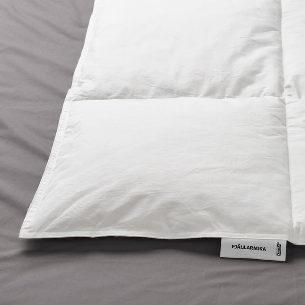 White duvet with stitched edges rests on grey bedsheets. White tag with FJÄLLARNIKA and ikea logo is visible at one end.