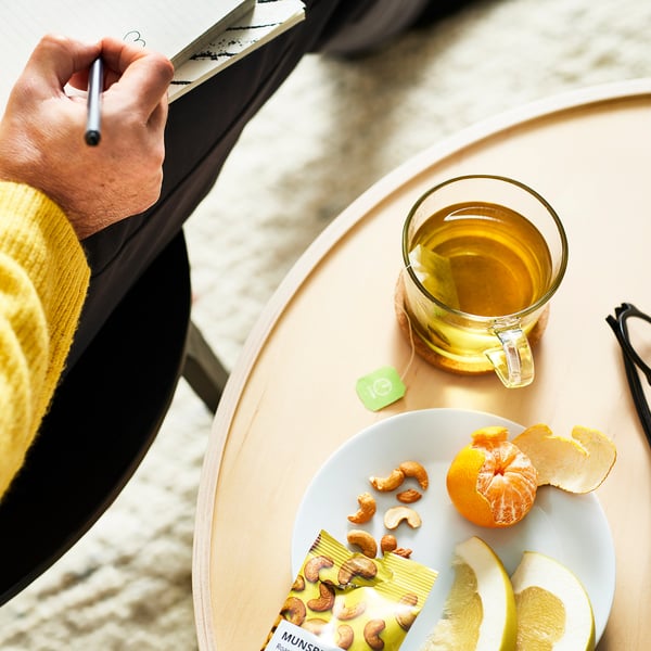 Person writing in notebook beside a table with tea, fruit, and cashew nuts.