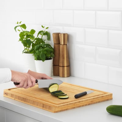 Person slicing cucumber on wooden cutting board with two knives beside potted herbs and wooden mills.