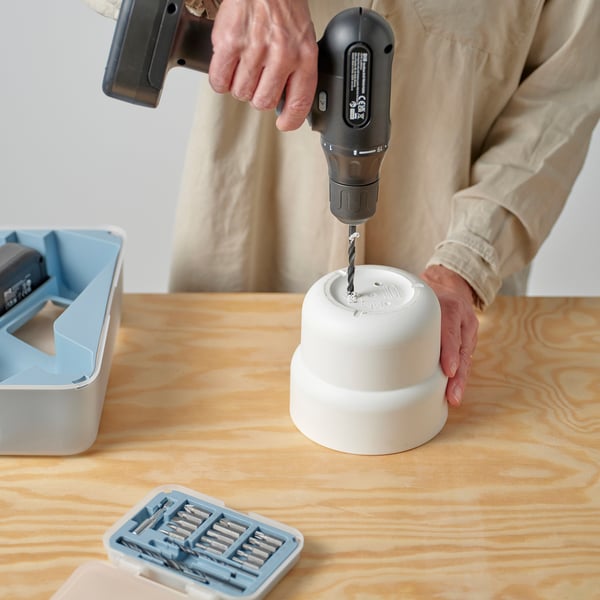 Person drilling a hole into a white planter on a wooden table, demonstrating the pots durability and ability to create drainage.
