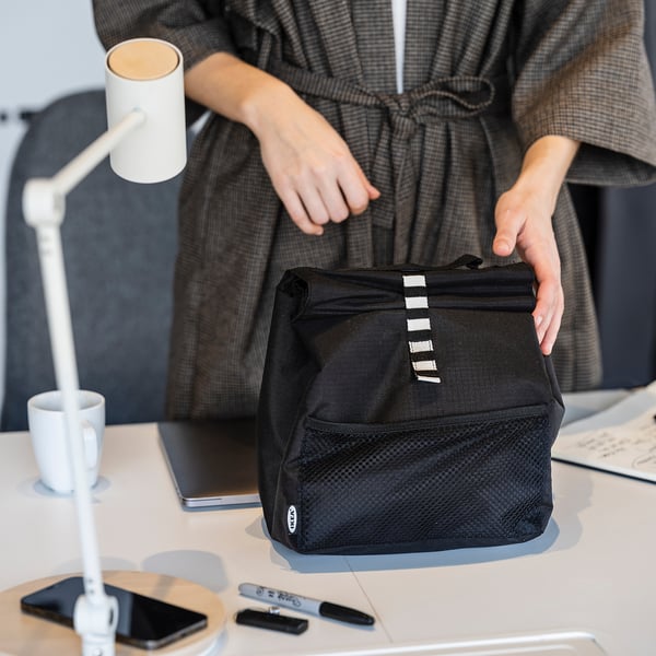 A person holding a black FRAMTUNG lunch bag on a desk with a laptop, phone, and mug.