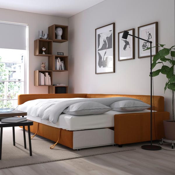 Modern sofa-bed with storage, shown as bed with white pillows and orange upholstery, next to a wooden bookshelf with decor.