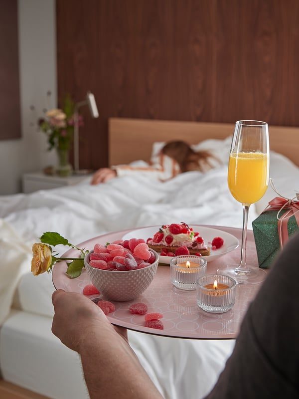 A person serves breakfast in bed, including pastries, candies, a glass of mimosa, candles, and a rose.