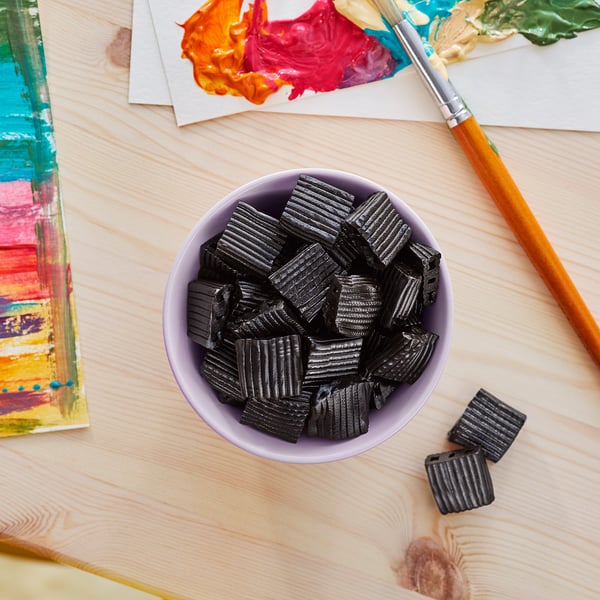A bowl of black licorice candies next to colourful paints and brushes on a wood table.