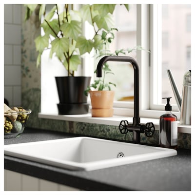 Black kitchen tap with two handles set in a white farmhouse sink on a grey countertop next to potted plants.