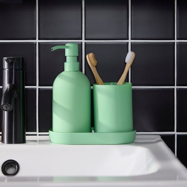 Green soap dispenser and toothbrush holder on white sink. Tray keeps items organised. Against dark tiled wall.