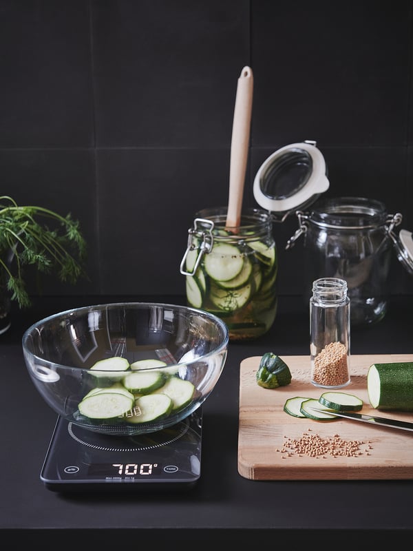 Kitchen setup: scale at 700g, sliced cucumbers, seeds, knife, pickles, spices, herbs, dark background.
