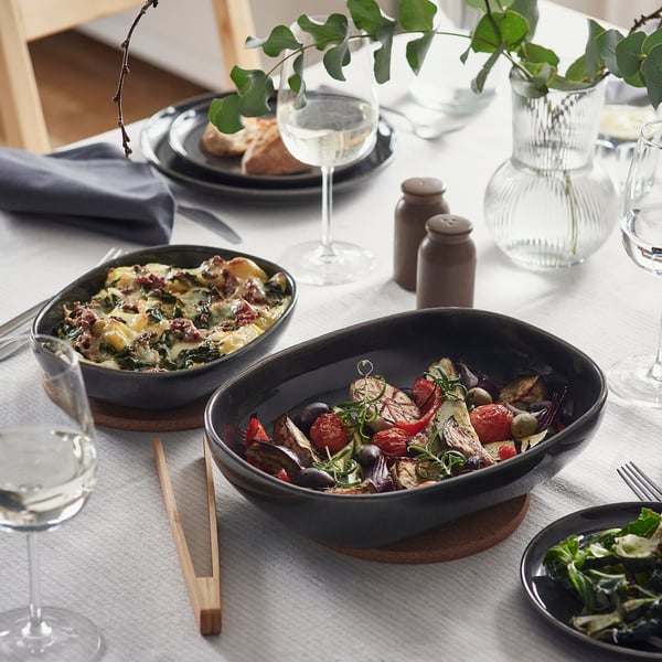 Two dark grey oven dishes on a table with various foods and glasses. One dish has baked food, the other roasted vegetables.