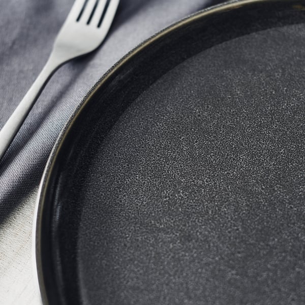 Black GLADELIG stoneware plate with textured glaze sits next to a fork on a dark cloth.