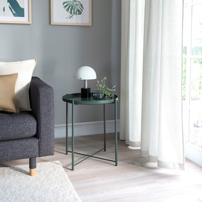 Small dark green side table with round tray top next to grey couch under white lamp.