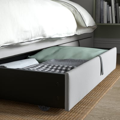 A low storage box under a bed, partially open, revealing neatly folded bedding items. The box is on wheels for easy access.