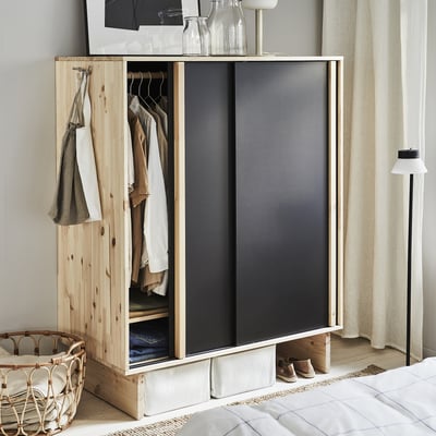 Natural wood and dark grey sliding wardrobe, open to display clothes, situated beside a bed with a woven basket and a floor lamp.