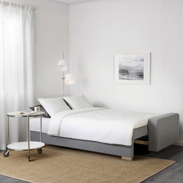 Minimalist bedroom with grey sofa-bed, white sheets, side table, lamp, and beach photo frame.