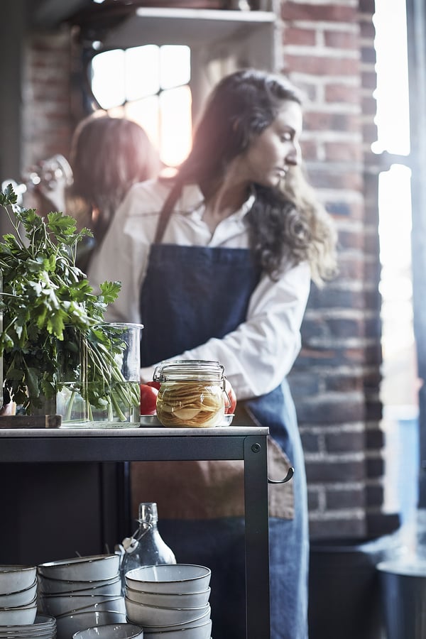 Person in kitchen, wearing blue apron, interacting with stainless steel table, holding jar.