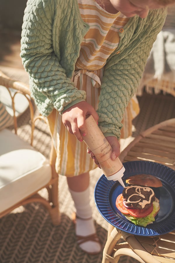 Person adds sauce from white GRILLTIDER bottle to burger on blue plate, seated outdoors.