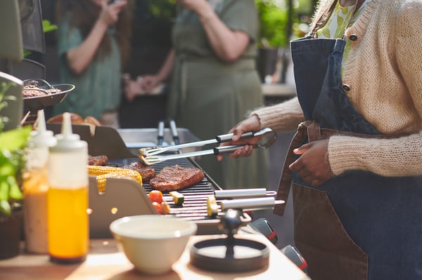 Outdoor grilling scene, person holding GRILLTIDER in hand, various condiments nearby, steak on grill.