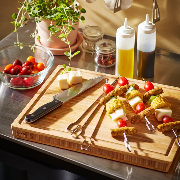 Wooden cutting board with knife, cheese, tomatoes, skewers, bowls with tomatoes, bottles with oil and vinegar, plant in background.