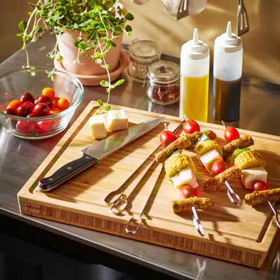 Wooden cutting board with knife, cheese, tomatoes, skewers, bowls with tomatoes, bottles with oil and vinegar, plant in background.