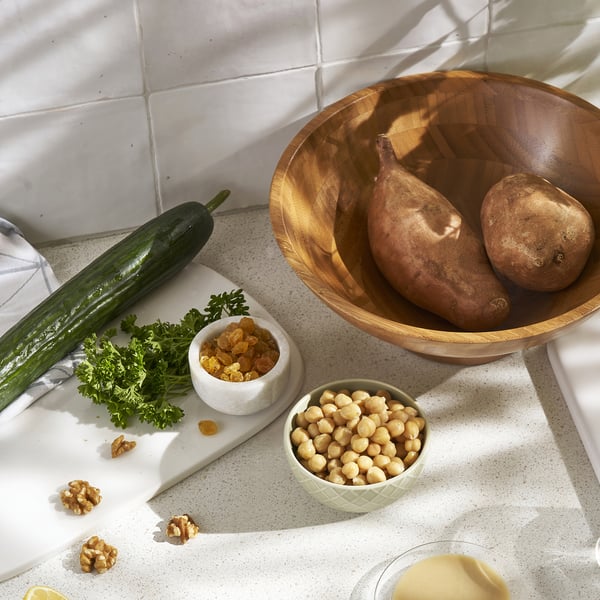 Wooden bowl, small dishes with vegetables, chickpeas, and nuts on a white speckled countertop, against a tiled wall.
