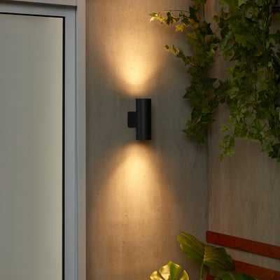 Black GRÖNSPRÖT wall lamp illuminates wall. Sleek, compact design; emitting warm light. Surrounded by plants; smaller than adjacent door.