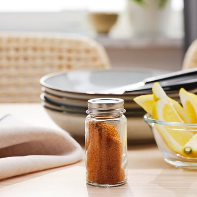 Small glass jar with spice, lid on, on table next to lemon slices, napkin, and plates.