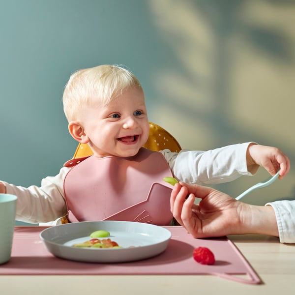 Baby wearing a silicone bib with a pocket catching dropped food. An adult is feeding the baby with a spoon. A bowl with fruit is on the table.
