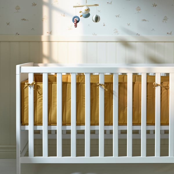 Image shows a baby crib with a padded bumper. The bumper, secured by ribbons, softens the hard slats and ensures safety.