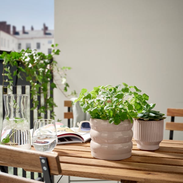 Pink HASSELBUKETT plant pots in stoneware with unique glaze variations hold green plants on a wooden table, accompanied by glasses and a magazine.